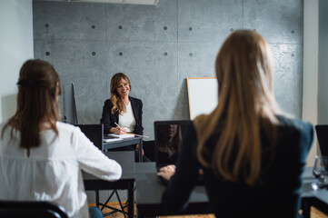 Cheerful,middle-aged caucasian lecturer takes notes while she listens to young female employees brainstorming in company.Two young participants have theirbacks turned. Whiteboard is beside the teacher