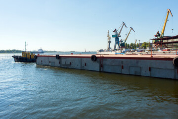 a river tug pulls a barge against the backdrop of shipyard cranes