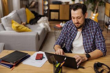 A man in a white T-shirt works alone at home typing out the latest documents. A phone with a payment card, documents and headphones lie next to him