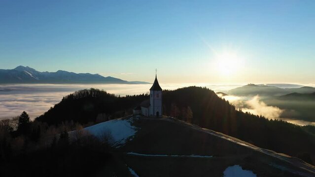 Church of St. Primoz and Felicijan at Sunrise above the Clouds. Julian Alps. Jamnik, Slovenia, Europe. Aerial View. Orbiting