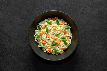 Instant noodles with vegetables in bowl, on dark background, top view, space to copy text.