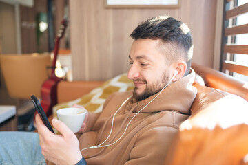 cheerful young man wearing headphones and holding smart phone with smile while sitting on the couch at cafe