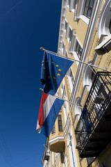 Flags of the Netherlands and the European Union hang from flagpoles against the blue sky and the facade of the building