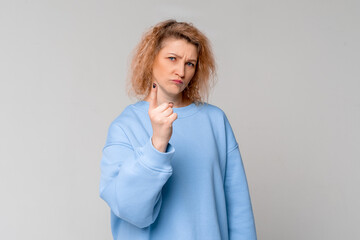 Serious middle age woman with curly blonde hair shakes her finger threateningly, scolding or telling off someone, standing in trendy blue sweatshirt over light grey background