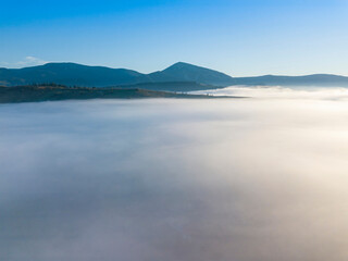 Flight over fog in Ukrainian Carpathians in summer. Mountains on the horizon. A thick layer of fog covers the mountains with a continuous carpet. Aerial drone view.