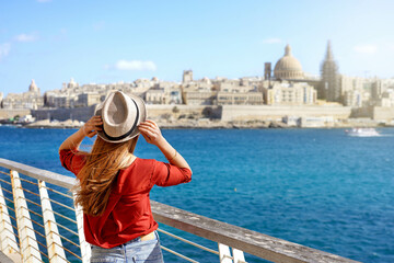 Tourism in Europe. Traveler girl holding hat walking along Malta promenade with Valletta cityscape on the background. - obrazy, fototapety, plakaty