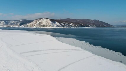 Drone is moving on the surface of Lake Baikal. The coastal part is covered with thin ice. On the opposite bank of the hills and the village at their foot. Cinematic calm landscape in summer clear day