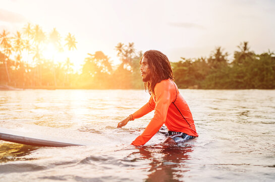 Black long-haired teen man floating on long surfboard, waiting for a wave ready for surfing with palm grove litted sunset rays. Extreme water sports and traveling to exotic countries concept.