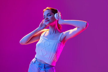 Portrait of young beautiful happy girl in headphones listening to music isolated on purple background in neon light, filter. Concept of emotions, music, facial expressions