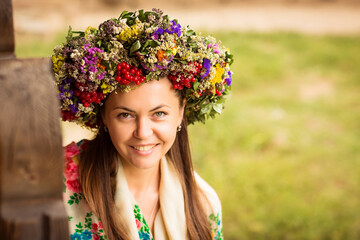 Young pretty ukrainian woman with a wreath of dry herbs and viburnum berries on her head sitting near a wooden house