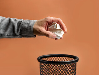 Man's hand throws crumpled paper into a basket on a beige background with copy space.
