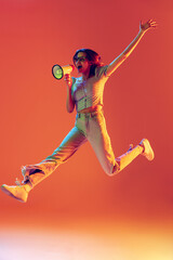 One slim beautiful excited girl jumping with megaphone isolated on peach color background in neon...