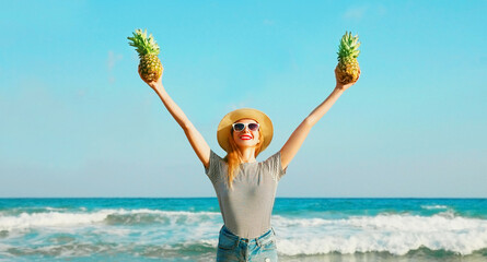 Happy smiling young woman raising her hands up with pineapples on the beach on sea background at summer day