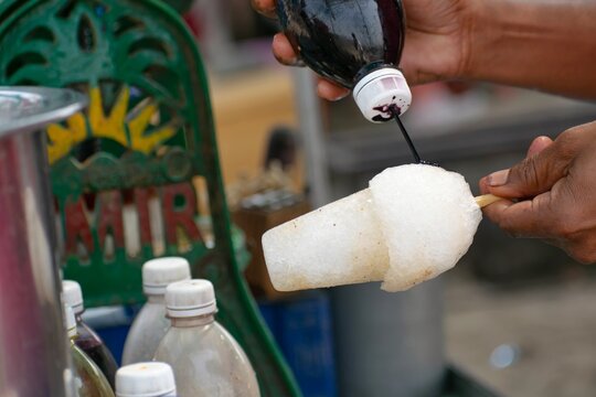 A picture of hands of a hawker holding ice gola (ice candy) and pouring kala khatta flavoured syrup on a hot sunny day for his customers at beach.