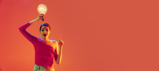 Fototapeta na wymiar Flyer with emotional young girl holding big electric bulb over her head isolated on peach color background in neon filter. Concept of emotions, beauty, fashion, youth