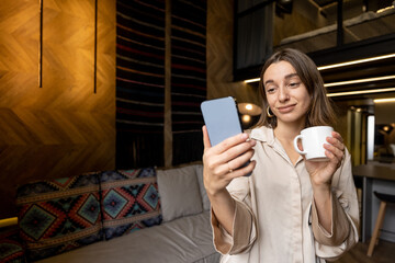 Woman using smart phone while standing with coffee cup in living room of a modern style apartment in the morning. Concept of online communication and home comfort