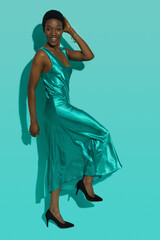 Smiling beautiful black woman in a turquoise satin long dress is standing on one leg.