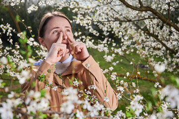 Woman allergic suffering from seasonal allergy at spring, posing in blossoming garden at...