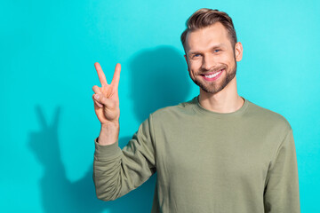 Photo of young blond funky guy show v-sign wear khaki pullover isolated on teal color background