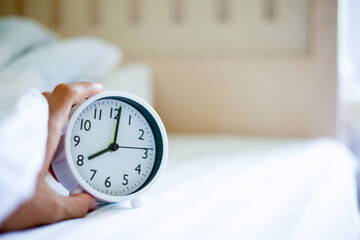 Lying under the covers, reaching for the alarm clock. A white clock near the bed showing morning time on the background of white bed linen. The concept of waking up early. space for text.