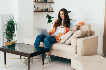 Young modern redhead woman sitting in casual clothes at home on the sofa uses a laptop.