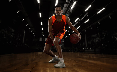 African-american young basketball player in action and motion in flashlights over dark gym...