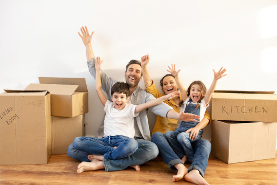 Happy multiracial family of four sitting on floor surrounded cardboard boxes in empty living room. Owners of estate, mortgage