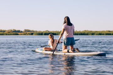 Mother and little daughter on sup board rowing together effortfully, exercising and having fun on lake with green reeds in background. Active lifestyle. Teaching to do sports from childhood.