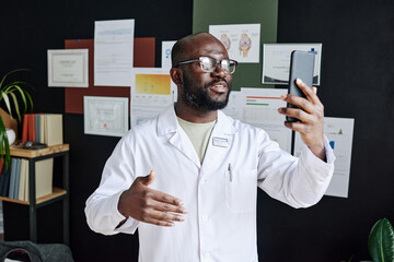 African male doctor in white coat using mobile phone for online consultation during work at office