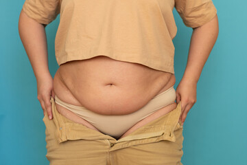 Unrecognizable plump plus-size overweight woman in beige T-shirt pulling on unzipped jeans, showing...