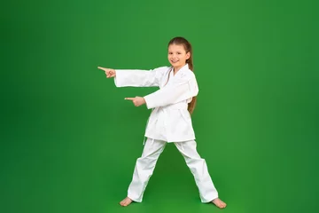 Zelfklevend Fotobehang A girl in a kimono on a green background points her fingers at the advertisement. kid studies martial arts. child development in sports. © Юлия Дьякова