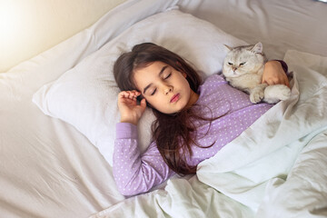 A little brunette girl in purple pajamas sleeps hugging a white charming cat lying on a white pillow on the bed.
