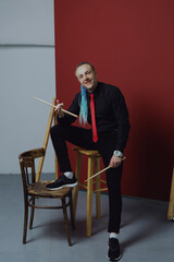 A young attractive man with an interesting hairstyle in pigtails on an undercut with a red tie on a red background in and drumsticks in his hands