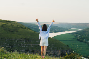 Fototapeta na wymiar Happy free woman with raised hands standing on a hill above the river at sunny summer day. Travel, freedom, happiness.