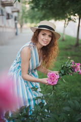 Fototapeta premium Portrait of young redhead curly woman in straw hat and linen stripe dress with a basket and a pink peonies bouquet in the garden