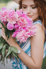 Portrait of young redhead curly woman in straw hat and linen stripe dress with a basket and a pink peonies bouquet in the garden. No face closeup