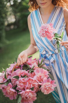 Portrait of young redhead curly woman in straw hat and linen stripe dress with a basket and a pink  peonies bouquet in the garden. No face closeup