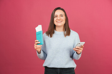 Excited woman has just checked in online on the phone while holding passport. Studio shot over pink wall.