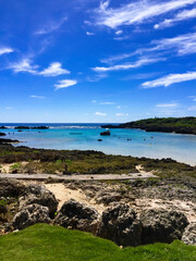 宮古島の真っ青なシギラビーチと青空と岩場の壮大な景色、そして遠くのボートパラソルと海水浴客 - obrazy, fototapety, plakaty