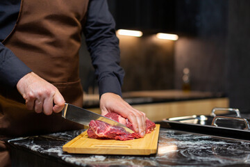 A butcher in an apron in the kitchen cuts pork on a wooden board. Steak.