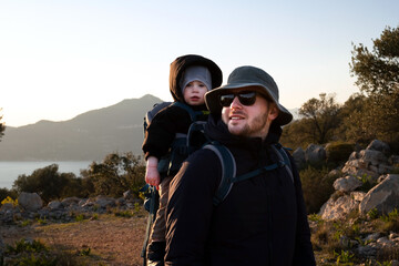Man wearing sunhat, sunglasses carrying baby,toddler in backpack.Young handsome father traveling,hiking with kid, child of 1,2 years old.Family trip in nature,mountains,cold weather near sea