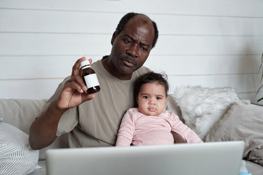 Modern Mature Black Man Sitting On Sofa In Front Of Laptop With Baby Daughter On His Lap Having Online Consultation With Pediatrician