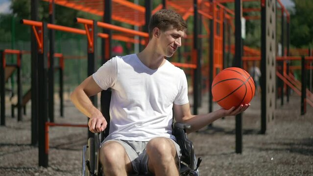 Young smiling man in wheelchair bouncing ball in slow motion and looking at camera. Portrait of confident inspired positive Caucasian person with disability posing in sunny park on sports ground