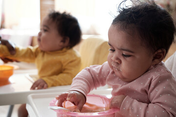 African American baby girl sitting in high chair touching fruit puree with hand during lunch time