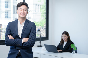 Asian young successful businessman wear formal suit smiling with confident and standing with crossed arm at office with his colleagues woman working on laptop on blur background. White background