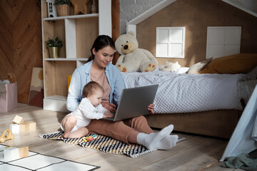 Modern young woman sitting on floor in kids room working in Internet on laptop and looking after her daughter