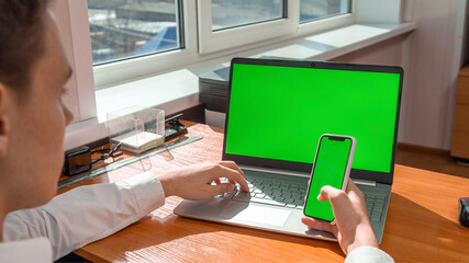 A young man works in the office behind a laptop with a green screen, in his hands holds a phone with a green screen. High quality photo