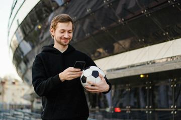 Smiling football fan with soccer ball holding smartphone and using betting app. Football stadium on the background. Copy space. Gambling concept.