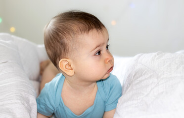 super cute baby boy lying on bed, on belly with serious face or cry out, nervous kid. visible first teeth, teething. white blanket and lights in the back. adorable boy for advertising, diapers or care