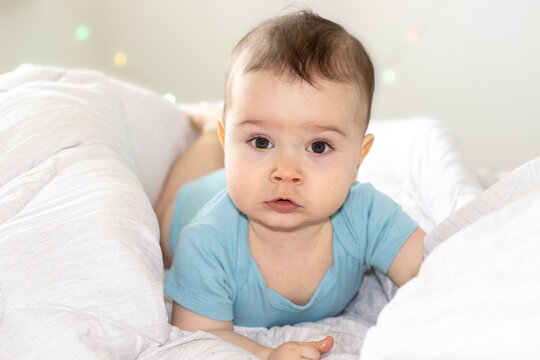 super cute baby boy lying on bed, on belly with serious face or cry out, nervous kid. visible first teeth, teething. white blanket and lights in the back. adorable boy for advertising, diapers or care